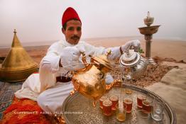 Image du Maroc Professionnelle de  Un marocain traditionnellement vêtu serve du thé à la menthe aux invités du grand Moussem de Tan Tan. Installée avec ses plateaux sur les hauteurs de la plage Chbika au environ de Tan Tan. Le Moussem est une sorte de foire annuelle, à la fois économique, culturelle et sociale. Il est classé par l'Unesco en 2005 comme patrimoine immatériel humain mondial. Chaque année la majorité des tribus et des grandes familles nomades du désert se retrouvent sur un site désertique pour célébrer ce grand Moussem où  danse, chants, course de chameaux et fantasia font partie des festivités. , Samedi 18 septembre 2004, ((Photo / Abdeljalil Bounhar) 